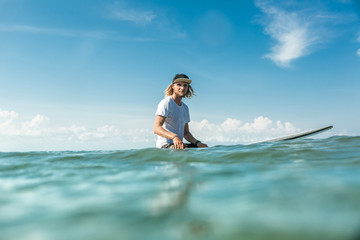 selective focus of young male surfer sitting on surfing board in ocean at Nusa Dua Beach, Bali, Indonesia
