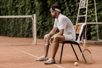 side view of handsome retro styled tennis player sitting on chair with bottle of water at tennis...