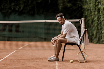 handsome retro styled tennis player sitting on chair with bottle of water at tennis court