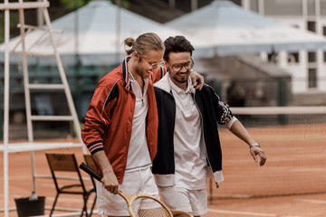 smiling retro styled tennis players walking with rackets and hugging at tennis court