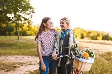 Obraz premium Two beautiful smiling girls with bicycle and basket full of wildflowers and fruits happily looking on each other while spending time in park