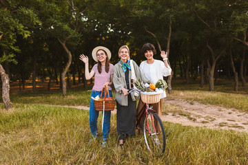 Group of happy girls with bicycle and baskets full of wildflowers and fruits joyfully waving and looking in camera in city park