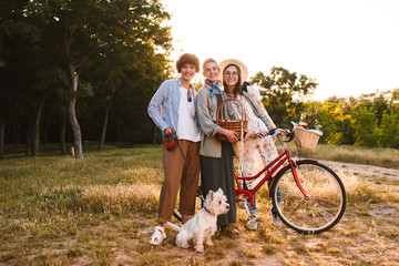 Pretty smiling girls with bicycle and wood basket in hands happily looking in camera while spending time with little dog in park