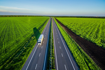 White trucks driving on asphalt road along the green fields in rural landscape at sunset. Road seen...