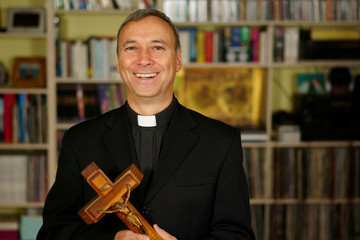 A good looking smiling and laughing catholic priest is showing a crucifix. He looks at us with interest, pensiveness, proselytising, joy and happyness. - obrazy, fototapety, plakaty