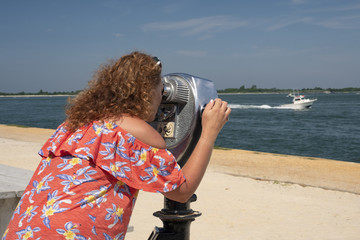 woman uses coin operated binocular to observe landscape, ocean, boats, beach scenes at the coast
