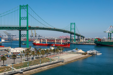 Red Bulk Carrier Ship Passing Under the Vincent Thomas Bridge and Container Ships Unloading in Los Angeles Long Beach California Shipping Port - obrazy, fototapety, plakaty