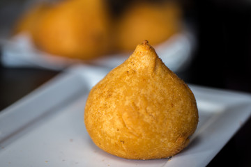 salgadinho frito servido em prato branco com  outros salgados desfocados no fundo