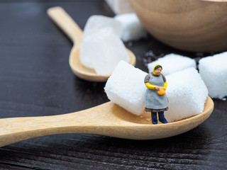 Miniature fat woman standing near wooden bowl and spoon with sugar cube on dark wood background, and thinking of weight loss and slim body. Healthy lifestyle concept.