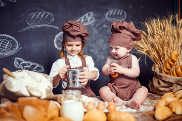 Little girl with baby in aprons on table with bread loaves making fresh dough and having fun