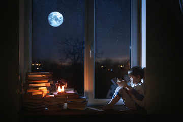 Fototapeta na wymiar A cute boy sits on the windowsill at night. The child reads books under the moonlight. The window shows the moon and stars.