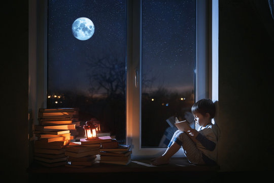 A cute boy sits on the windowsill at night. The child reads books under the moonlight. The window shows the moon and stars.