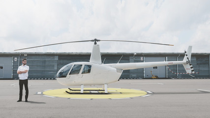 Portrait of commercial pilot in uniform standing near small private helicopter on a landing point © supamotion