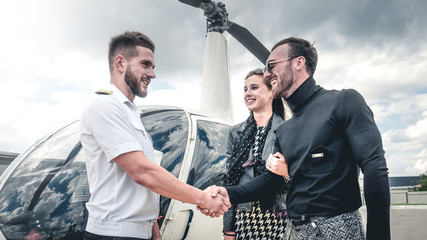 Commercial pilot in uniform greeting clients near small private helicopter on a landing point