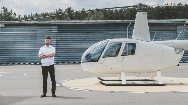 Portrait of commercial pilot in uniform standing near small private helicopter on a landing point