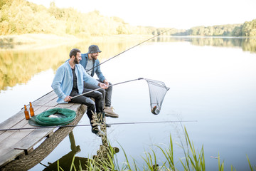 Two friends catching fish with fishing net and rod sitting on the wooden pier at the lake