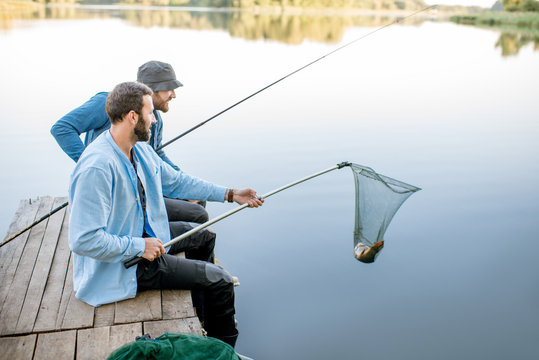 Two friends catching fish with fishing net and rod sitting on the wooden pier at the lake