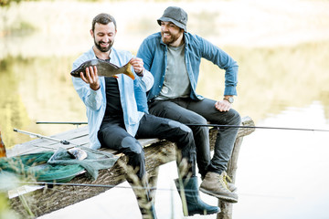 Two happy fishermen holding caught fish sitting on the wooden pier during the fishing on the lake...