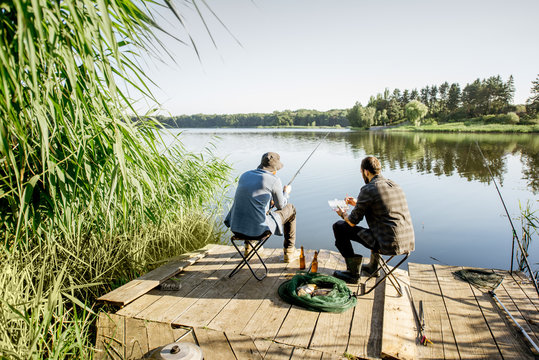 Landscape view on the beautiful lake and green reeds with two men fishing on the wooden pier during the morning light