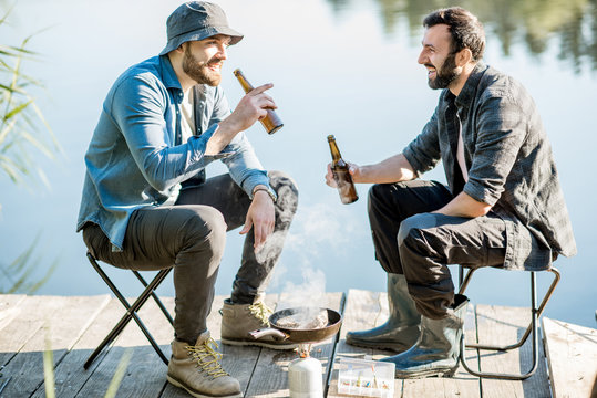 Two fishermen frying fish sitting with beer during the picnic on the wooden pier near the lake in the morning