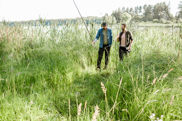 Two fishermen walking with fishing rod and net on the green lawn near the lake in the morning