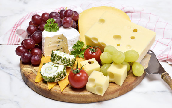 Delicious cheese plate with various sorts of cheese like Emmentaler, gouda and brie. Gourmet cheese on a wooden cutting board with white marble background and kitchen towel.