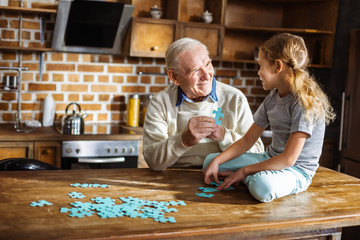 Cheerful aged man assembling pazzles with his granddaughter