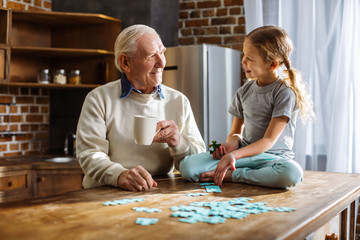 Joyful aged man assembling jigsaw puzzles with his granddaughter - obrazy, fototapety, plakaty