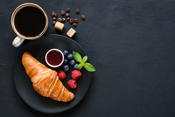 Croissant, fresh berries, jam and cup of black coffee on black background. Top view, copy space for...