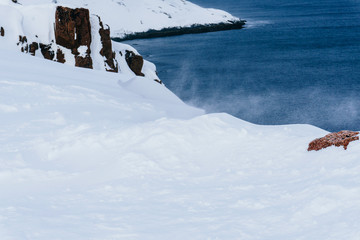 High red rocks rise above the snow-covered coast of the sea in winter in spooky cold and strong wind during an expedition to the Arctic