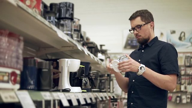 Adult man is rotating a detail of electrical coffee-machine in a trading floor of a shop. He is putting it on a shelf and getting coffee-maker
