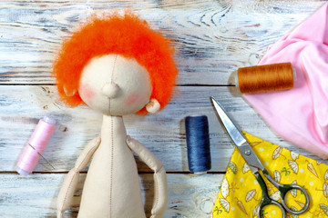 Sewing and needlecraft art concept.Scissors, spools of thread preparation for body of textile doll boy with ginger hair, on white wooden background. Colorful threads and needles for handicraft.