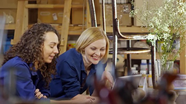 Two attractive Caucasian women in workwear leaning on table and using laptop computer in carpentry workshop