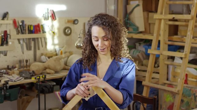 Medium shot of friendly curly haired woman speaking and polishing wooden frame with sandpaper in carpentry workshop