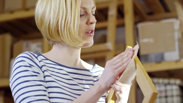 Tilt up shot of attractive young woman polishing wooden frame and talking to someone in carpentry workshop