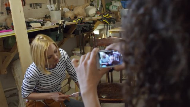 Female artisan applying varnish on old wooden chair and talking to camera while her friend is holding smartphone