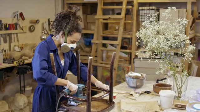 Curly haired woman in workwear and respirator using sanding machine to polish surface of old wooden chair in carpentry workshop