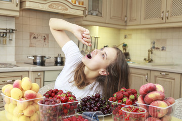 Beautiful young girl eats fruits and fresh berries in the kitchen, beautifully lays cherries in the mouth