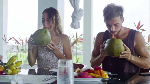 Medium shot of Caucasian couple sitting at table on balcony and drinking coconut water when enjoying healthy breakfast