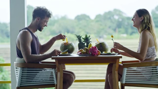 Side view of happy Caucasian couple sitting opposite each other at table on balcony, laughing and talking to each other when enjoying healthy breakfast of tropical fruits