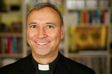 A portrait of a good looking smiling catholic priest in his library. He looks at us with interest,...