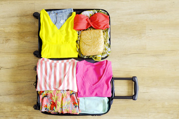 Open suitcase fully packed with folded women's clothing and accessories on the floor. Woman packing for tropical vacation concept. Female luggage w/ things. Background, close up, copy space, top view.