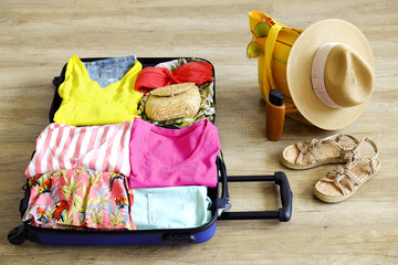 Open suitcase fully packed with folded women's clothing and accessories on the floor. Woman packing for tropical vacation concept. Female luggage w/ things. Background, close up, copy space, top view.