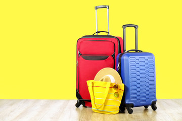 Red textile suitcase & blue hard shell luggage, extended telescopic handle, straw hat, yellow beach bag, mirrored sunglasses, yellow wall background. Couples retreat trip concept. Close up, copy space