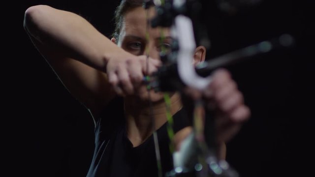Studio shot with tilt up of determined sportswoman shooting arrow with bow against black background