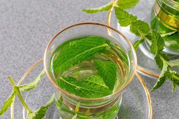 Close up view on a small tea glass filled with fresh peppermint tea beside green harvested peppermint plants on a grey table