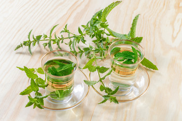 Two glasses with fresh peppermint tea in front of harvested green peppermint plants on a wooden table