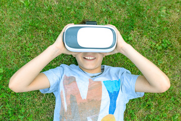 Young boy enjoying VR virtual reality outdoors. Caucasian pre teen lie down in the grass playing with VR. Dreaming of being part of a fantastic world thanks to new digital technologies.