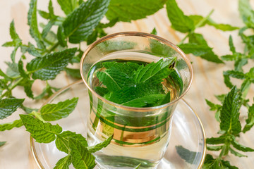 Close up view on a small tea glass filled with fresh peppermint tea beside green harvested peppermint plants on a wooden table