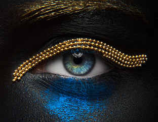 Macro and close-up creative make-up theme: beautiful female eyes with black skin with a gold chain and blue pigment